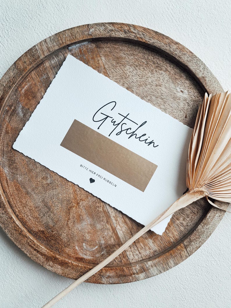May include: A white card with the word "Gutschein" written in black cursive. The card has a gold rectangle with the text "Bitte hier frei rubbeln" written in black below it.