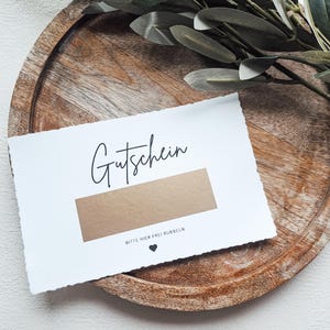 May include: A white card with the word "Gutschein" written in black cursive. The card has a brown scratch-off area with the text "Bitte hier frei rubbeln" (Please rub here freely) and a small black heart.