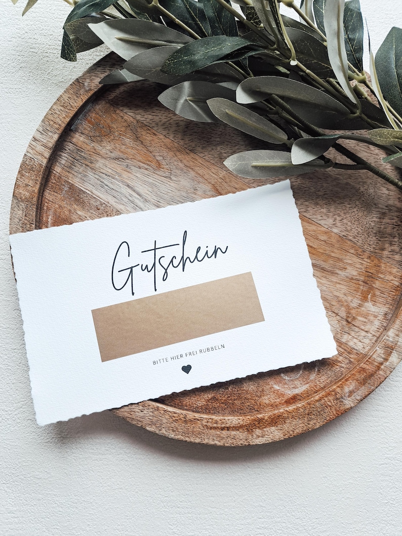 May include: A white card with the word "Gutschein" written in black cursive. The card has a brown scratch-off area with the text "Bitte hier frei rubbeln" (Please rub here freely) and a small black heart.