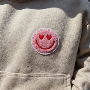 Könnte beinhalten: Beiger Kapuzenpullover mit einem rosafarbenen Smiley-Aufnäher. Der Aufnäher zeigt einen rosafarbenen Smiley mit herzförmigen Augen und einem roten Lächeln, umgeben von einem weißen Rand. Der Hoodie hat eine Kängurutasche.