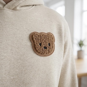 Könnte beinhalten: Beige Kapuzenpullover mit einem braunen Teddybär-Aufnäher auf der Brust. Der Teddybär-Aufnäher hat ein strukturiertes, gesticktes Aussehen mit einem braunen Rand und schwarzen Augen und Nase. Der Hoodie hat eine Kapuze mit Kordelzug.