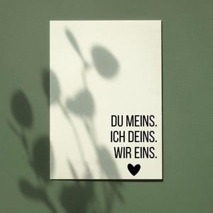 May include: A white rectangular card with the text "DU MEINS. ICH DEINS. WIR EINS." in black, with a black heart symbol. The card is set against a green background with plant shadows.