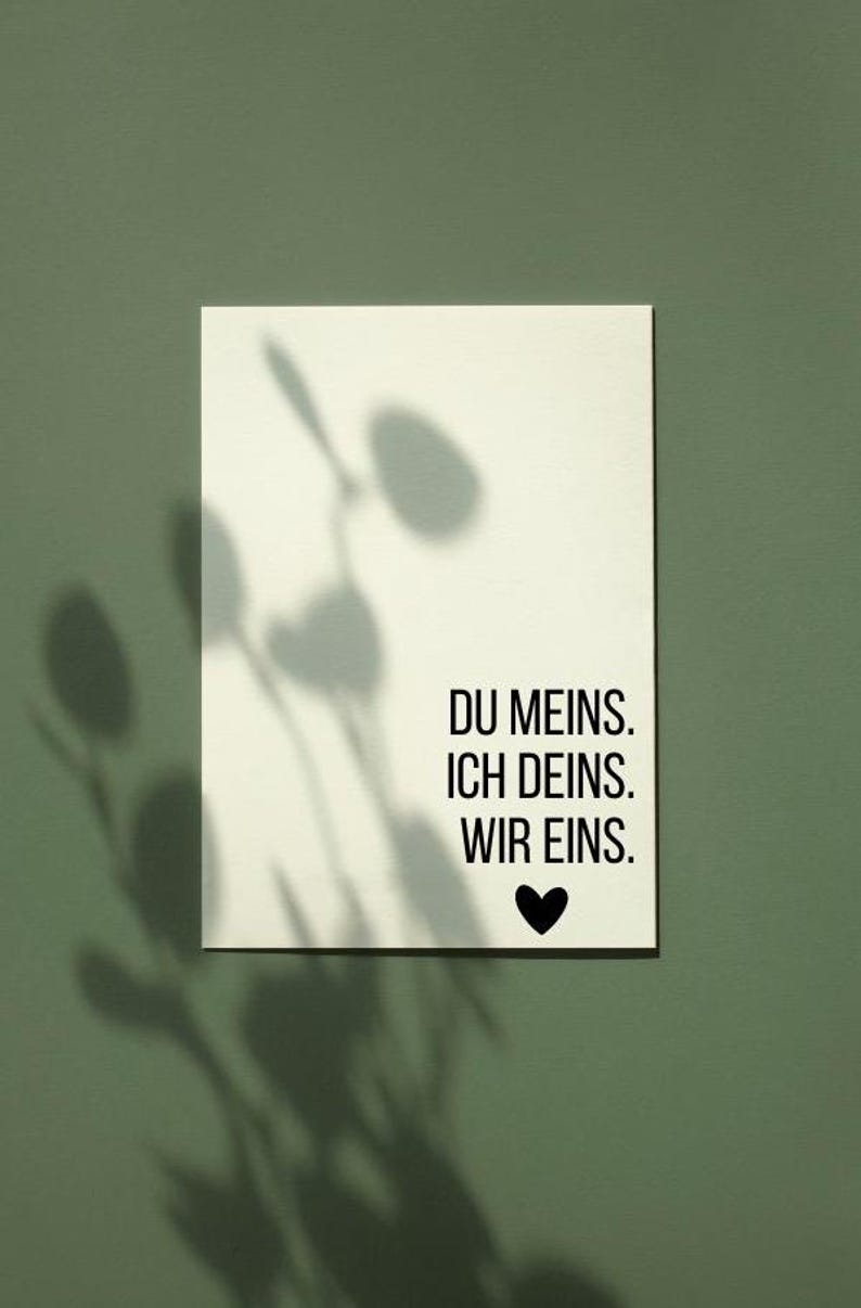 May include: A white rectangular card with the text "DU MEINS. ICH DEINS. WIR EINS." in black, with a black heart symbol. The card is set against a green background with plant shadows.