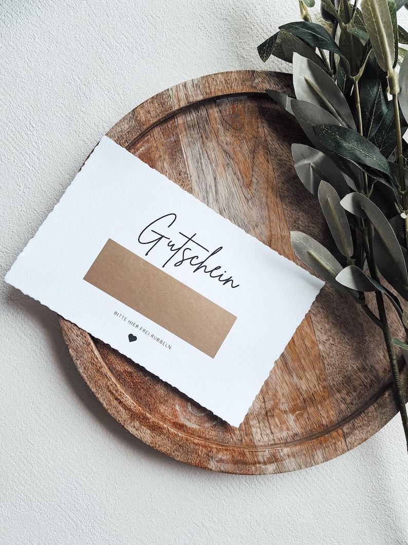 May include: A white card with the word "Gutschein" written in black cursive. The card has a brown scratch-off area with the text "Bitte hier frei rubbeln" and a small black heart.