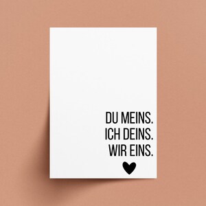May include: A white card with black text that reads "DU MEINS. ICH DEINS. WIR EINS." A black heart is below the text. The card is on a peach-colored background.