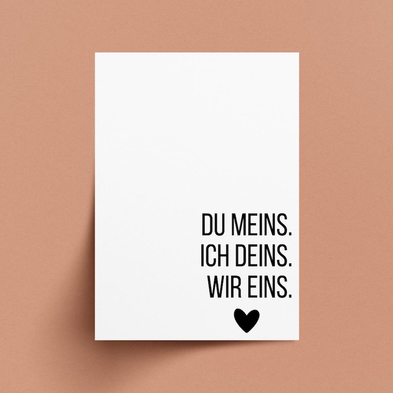 May include: A white card with black text that reads "DU MEINS. ICH DEINS. WIR EINS." A black heart is below the text. The card is on a peach-colored background.