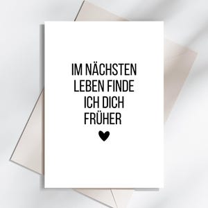 May include: A white greeting card with black text that reads "IM N&Auml;CHSTEN LEBEN FINDE ICH DICH FR&Uuml;HER" and a black heart. The card is on top of a white envelope, with a light gray background.