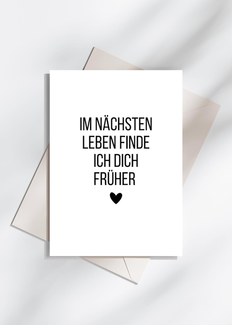 May include: A white greeting card with black text that reads "IM N&Auml;CHSTEN LEBEN FINDE ICH DICH FR&Uuml;HER" and a black heart. The card is on top of a white envelope, with a light gray background.