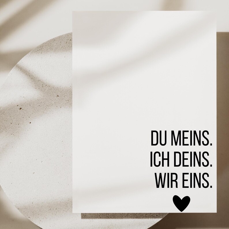 May include: A white card with the text "DU MEINS. ICH DEINS. WIR EINS." in black, accompanied by a black heart symbol. The card is set against a neutral background, with soft shadows.