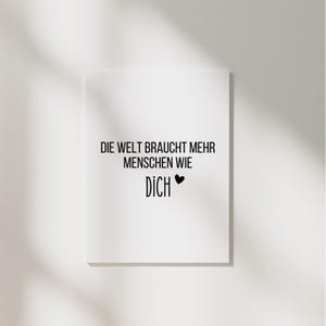 Könnte beinhalten: Ein weißes rechteckiges Schild mit schwarzem Text, der lautet: "DIE WELT BRAUCHT MEHR MENSCHEN WIE DICH" mit einem schwarzen Herzsymbol. Das Schild ist an einer weißen Wand befestigt.