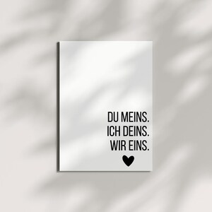 May include: A white rectangular print with black text that reads "DU MEINS. ICH DEINS. WIR EINS." A black heart is below the text. The print is on a white surface with shadows.