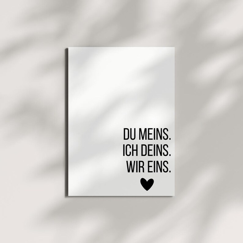 May include: A white rectangular print with black text that reads "DU MEINS. ICH DEINS. WIR EINS." A black heart is below the text. The print is on a white surface with shadows.