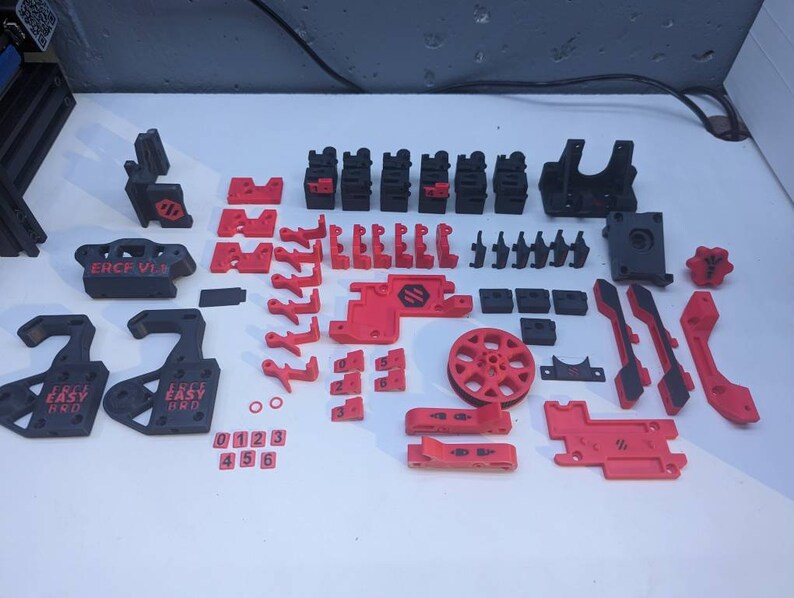 May include: A collection of 3D printed parts in black and red. The parts include a variety of shapes and sizes, including gears, wheels, and brackets. The parts are labeled with the text "ERCF V1.8" and "Easy Pro".