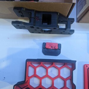May include: A 3D printed black and red plastic part with a honeycomb design. The part is made up of several pieces that fit together.