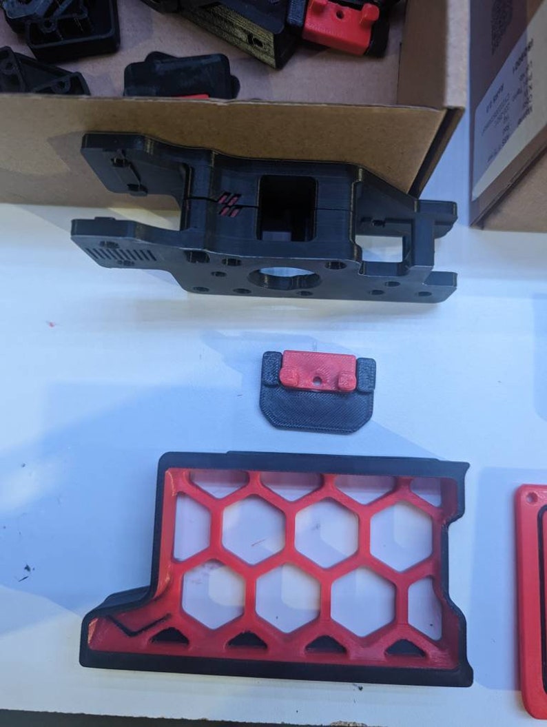 May include: A 3D printed black and red plastic part with a honeycomb design. The part is made up of several pieces that fit together.
