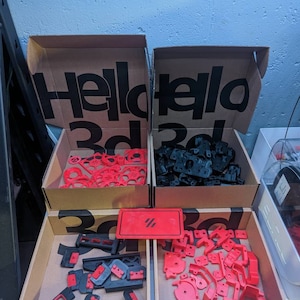 May include: Three cardboard boxes with the text "Hello 3d" printed on the top. The boxes are filled with red and black plastic parts. The boxes are sitting on a table.