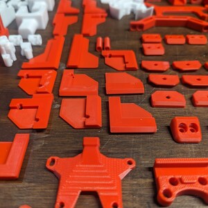 Puede incluir: Una colecci&oacute;n de piezas de pl&aacute;stico rojo impresas en 3D, que incluyen varias formas y tama&ntilde;os, como esquinas, conectores y una pieza en forma de estrella. Las piezas est&aacute;n dispuestas sobre una superficie de madera.