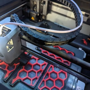 May include: Close-up of a 3D printer with a white cable running through a black chain. The printer bed is visible with a red honeycomb pattern and the text "Calibration Board" printed on the side.