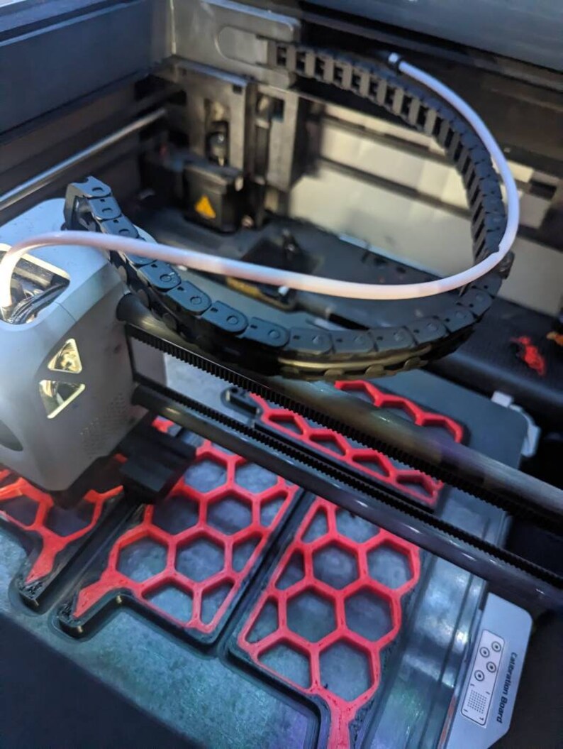 May include: Close-up of a 3D printer with a white cable running through a black chain. The printer bed is visible with a red honeycomb pattern and the text "Calibration Board" printed on the side.