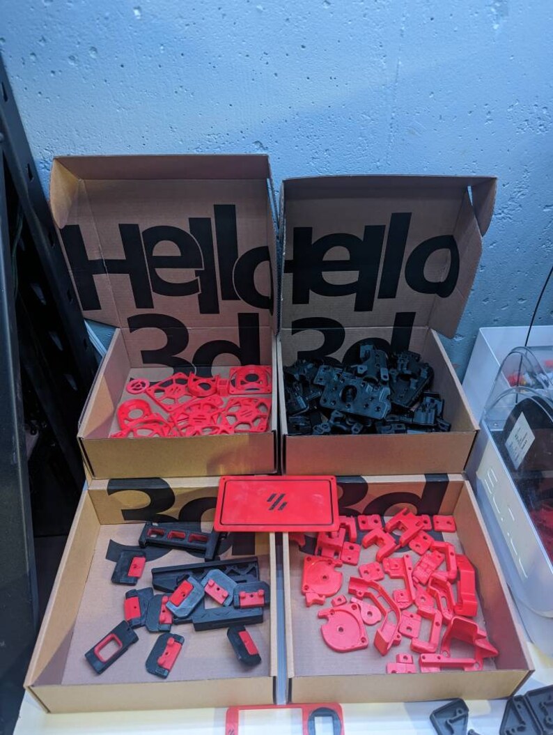 May include: Two cardboard boxes with the text "Hello 3d" printed on them. The boxes are filled with red and black plastic parts. The boxes are sitting on a table with a red sign that says "#" on it.