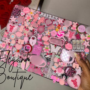 Könnte beinhalten: Ein rechteckiges Etui, bedeckt mit einer dichten Collage aus rosa und weißen dekorativen Anhängern, darunter Sterne, Herzen, Schleifen, Blumen, ein Parfümflakon und ein Schmetterling. Die Texte FEISTY, TAURUS und I LOVE ME sind zwischen den Objekten sichtbar.