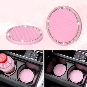May include: Two pink car cup holders with a diamond-like trim. The cup holders are shown in a car's cupholder area.