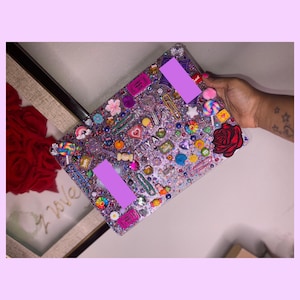 May include: A purple and silver glitter iPad case with a variety of colorful embellishments, including hearts, stars, flowers, and candy. The case has two purple rectangular stickers with the text "Liya" on one of them.