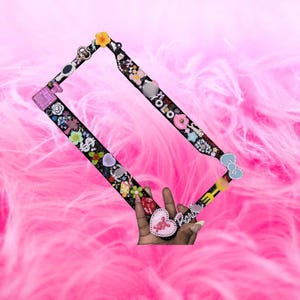 Puede incluir: Marco rectangular negro decorado con varios abalorios, incluyendo flores, corazones y la palabra "YOLO". El marco es sostenido por una mano contra un fondo rosa y plumoso. Los abalorios son de varios colores.