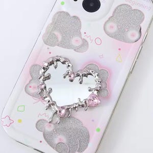 Può includere: Una custodia per telefono bianca con un design glitter rosa e argento. La custodia presenta uno specchio a forma di cuore argentato con strass rosa.