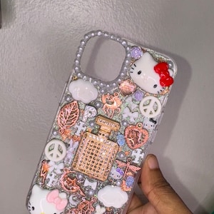 May include: A clear phone case with a glittery background and various charms, including a peace sign, a cloud, a butterfly, a Hello Kitty character, and a bottle of perfume. The case is decorated with rhinestones and pearls.