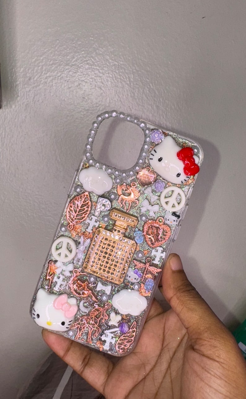 Puede incluir: Una funda transparente para tel&eacute;fono con fondo brillante y varios abalorios, incluyendo un signo de paz, una nube, una mariposa, un personaje de Hello Kitty y una botella de perfume. La funda est&aacute; decorada con diamantes de imitaci&oacute;n y perlas.