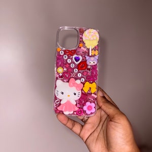 Puede incluir: Una funda de tel&eacute;fono rosa y blanca de Hello Kitty con purpurina y varios encantos, incluyendo una piruleta, un coraz&oacute;n, un lazo y una flor.
