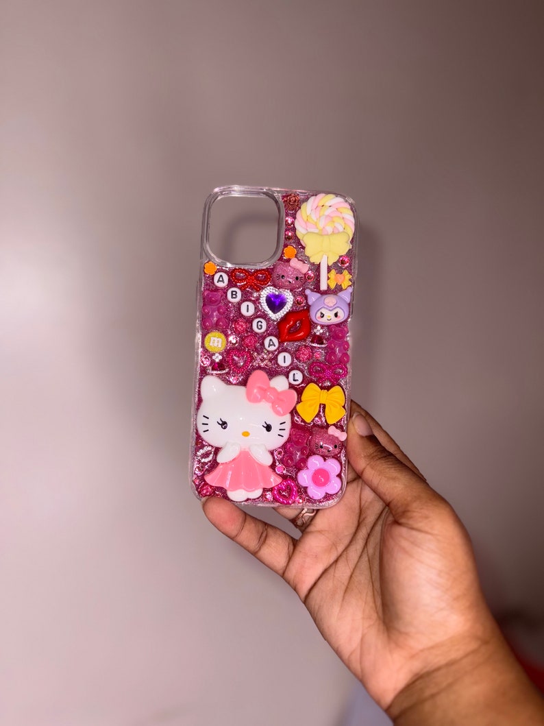 Puede incluir: Una funda de tel&eacute;fono rosa y blanca de Hello Kitty con purpurina y varios encantos, incluyendo una piruleta, un coraz&oacute;n, un lazo y una flor.