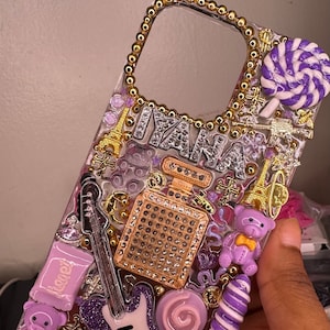 Puede incluir: Una funda transparente para tel&eacute;fono decorada con adornos morados, dorados y blancos. La funda presenta una guitarra morada, un osito de peluche morado, una piruleta morada y blanca y un frasco de perfume dorado con pedrer&iacute;a. La funda tambi&eacute;n tiene la palabra "IANA" escrita en pedrer&iacute;a plateada.