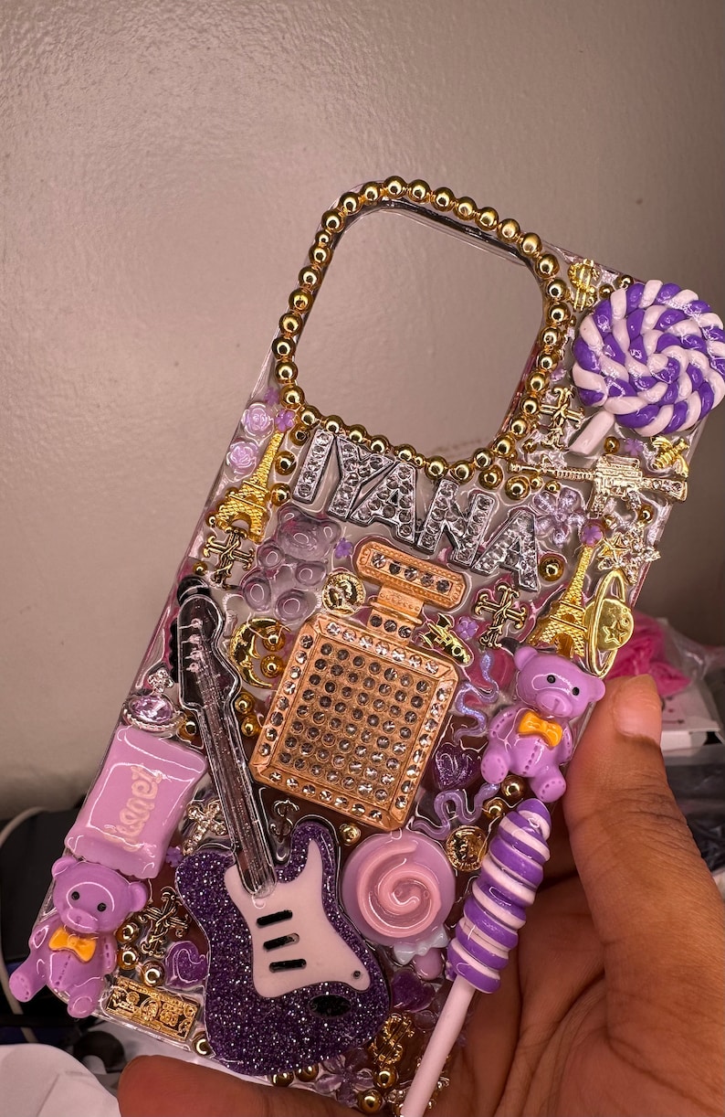 Puede incluir: Una funda transparente para tel&eacute;fono decorada con adornos morados, dorados y blancos. La funda presenta una guitarra morada, un osito de peluche morado, una piruleta morada y blanca y un frasco de perfume dorado con pedrer&iacute;a. La funda tambi&eacute;n tiene la palabra "IANA" escrita en pedrer&iacute;a plateada.