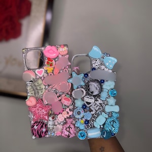 May include: Two clear phone cases decorated with various charms. The left case features pink and white accents, including a pink star, lips, a cupcake, and miniature money. The right case features blue accents, including a bow, a star, and the name JEN.