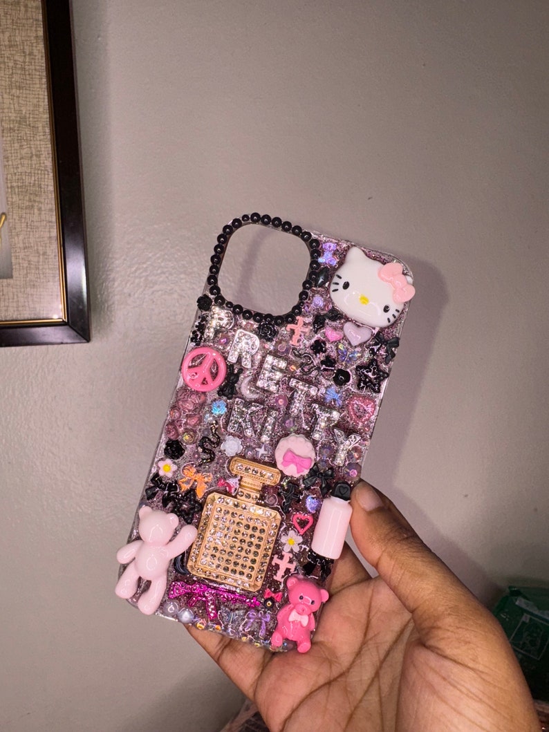 Puede incluir: Una funda de tel&eacute;fono rosa y negra con pedrer&iacute;a, purpurina y abalorios. La funda presenta un personaje de Hello Kitty, un signo de paz, un frasco de perfume y ositos de peluche.