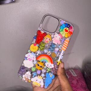 Puede incluir: Una funda transparente para tel&eacute;fono decorada con un arco&iacute;ris, una flor y varios personajes de dibujos animados. La funda est&aacute; adornada con purpurina y pedrer&iacute;a.