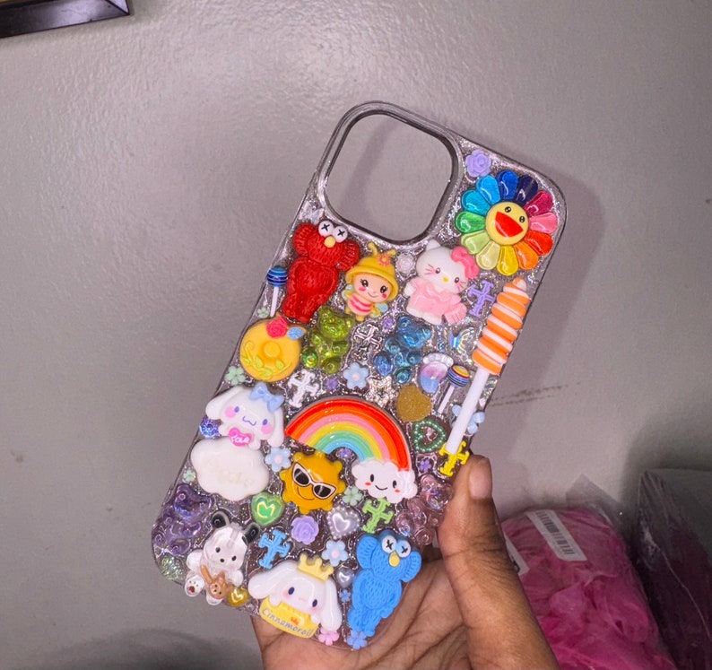 Puede incluir: Una funda transparente para tel&eacute;fono decorada con un arco&iacute;ris, una flor y varios personajes de dibujos animados. La funda est&aacute; adornada con purpurina y pedrer&iacute;a.