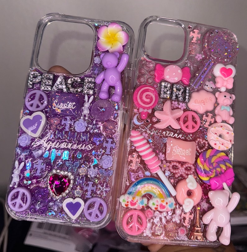 Puede incluir: Dos fundas transparentes para tel&eacute;fono decoradas con purpurina, abalorios y pedrer&iacute;a. Una funda es morada con las palabras "Peace" y "Aquarius" y la otra es rosa con la palabra "Bri" y abalorios con tem&aacute;tica de dulces.