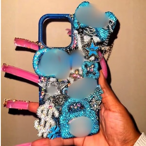 Puede incluir: Una funda de teléfono azul adornada con pedrería, estrellas y un signo de dólar. La funda está sujeta por una mano con uñas largas y rosas. La funda es un artículo decorativo y tiene una cadena.
