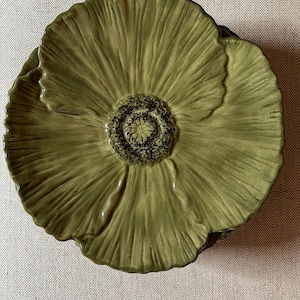 May include: A ceramic plate in the shape of a poppy flower. The plate is olive green with a textured surface, and the center has a dark brown, textured design. The plate is a decorative item.