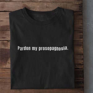 May include: Black t-shirt with white text that reads "Pardon my prosopagnosia."