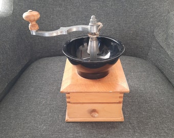 May include: A vintage wooden coffee grinder with a black ceramic bowl and a silver crank handle. The grinder has a small drawer on the bottom.