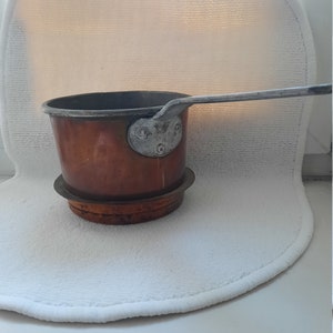 Könnte beinhalten: Ein kleiner Kupfertopf mit einem Metallgriff und einem Kupferboden. Der Topf steht auf einer weißen Oberfläche.