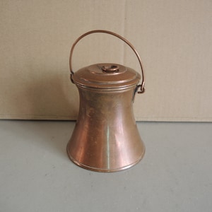 Puede incluir: Una pequeña jarra de cobre con asa y tapa. La jarra tiene forma de campana y una superficie lisa y brillante.
