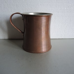 May include: A copper mug with a silver lining and a handle. The mug is on a white surface.