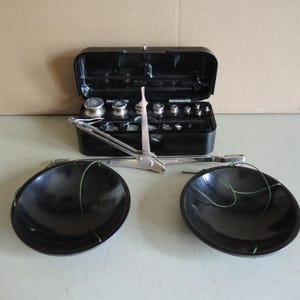 Op de afbeelding: Een vintage zwarte balansweegschaal met twee zwarte schalen en een set gewichten in een zwarte koffer. De weegschaal heeft een zilveren arm en een kleine zilveren aanwijzer. De gewichten zijn van verschillende maten en vormen.