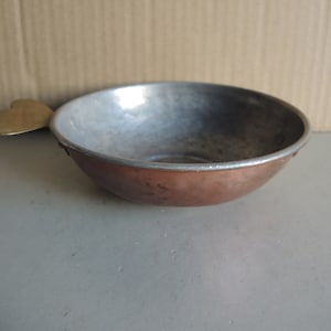 May include: A small, hammered copper bowl with a silver-coloured rim and a brass handle shaped like a stylized leaf. The bowl has a rounded shape and a tarnished appearance, suggesting age and use. The interior is a lighter, silver colour.