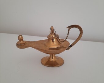 May include: A gold-colored genie lamp with a decorative handle and a chain. The lamp is on a white surface.